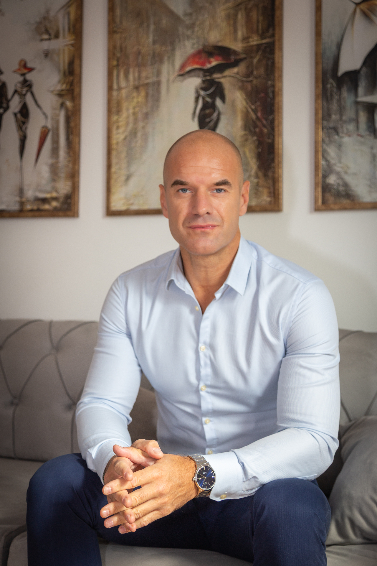 Nedeljko Rvović — seated calmly on a sofa in a light blue shirt, hands gently together, with artwork on the wall behind him